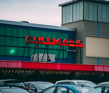 A Cinemark theater with a red neon sign, large windows, and a parking lot filled with cars in front.
